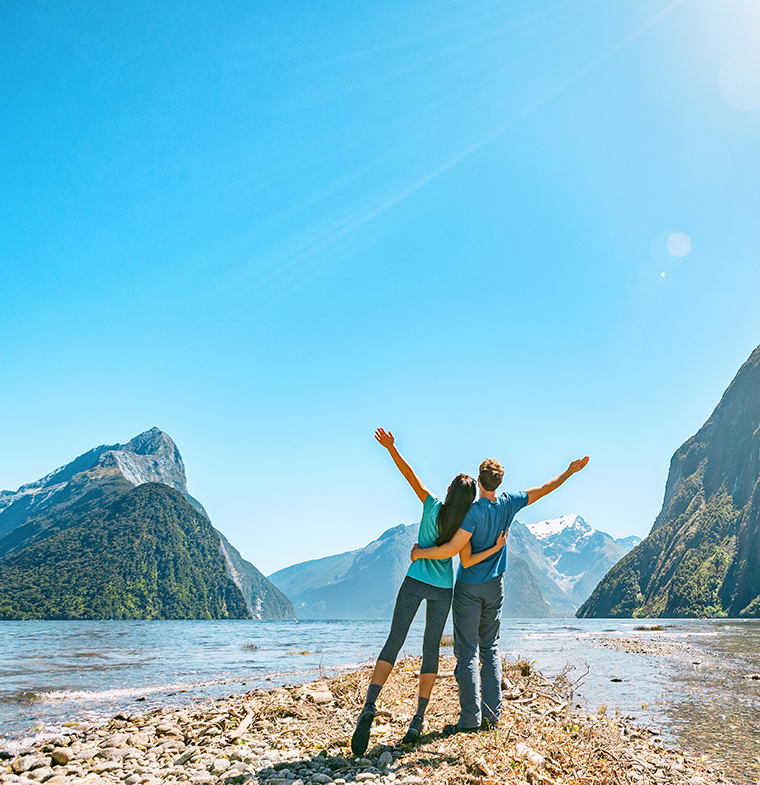 The Best Time to Visit Milford Sound Outdoor couple happy with arms outstretched in Milford Sound New Zealand in nature enjoying active outdoor lifestyle hiking in Milford Sound New Zealand by Mitre Peak in Fiordland